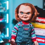 supreme scary chucky doll wearing outfit stands next colorful clothing indoors